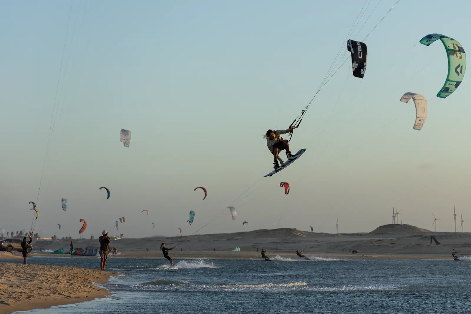 Kiter in salto freestyle con tavola e boots su una laguna piatta al tramonto, con decine di kite in volo, dune e turbine eoliche sullo sfondo