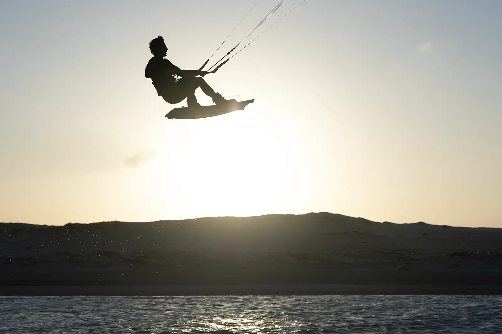 Kiter in silhouette in salto freestyle con tavola e boots, in controluce al tramonto sopra una laguna, con le dune sullo sfondo